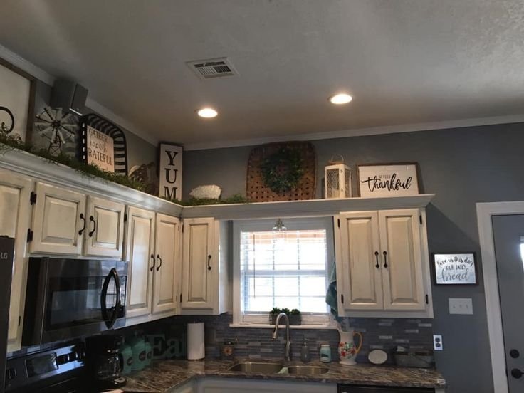 Above-cabinet decor styling with a woven basket, lantern and framed signs above white kitchen cabinets near a window