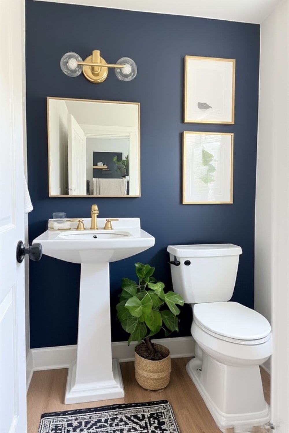 Deep navy blue powder room wall with a gold mirror, gold sconce, framed art and a fiddle leaf fig plant beside the toilet