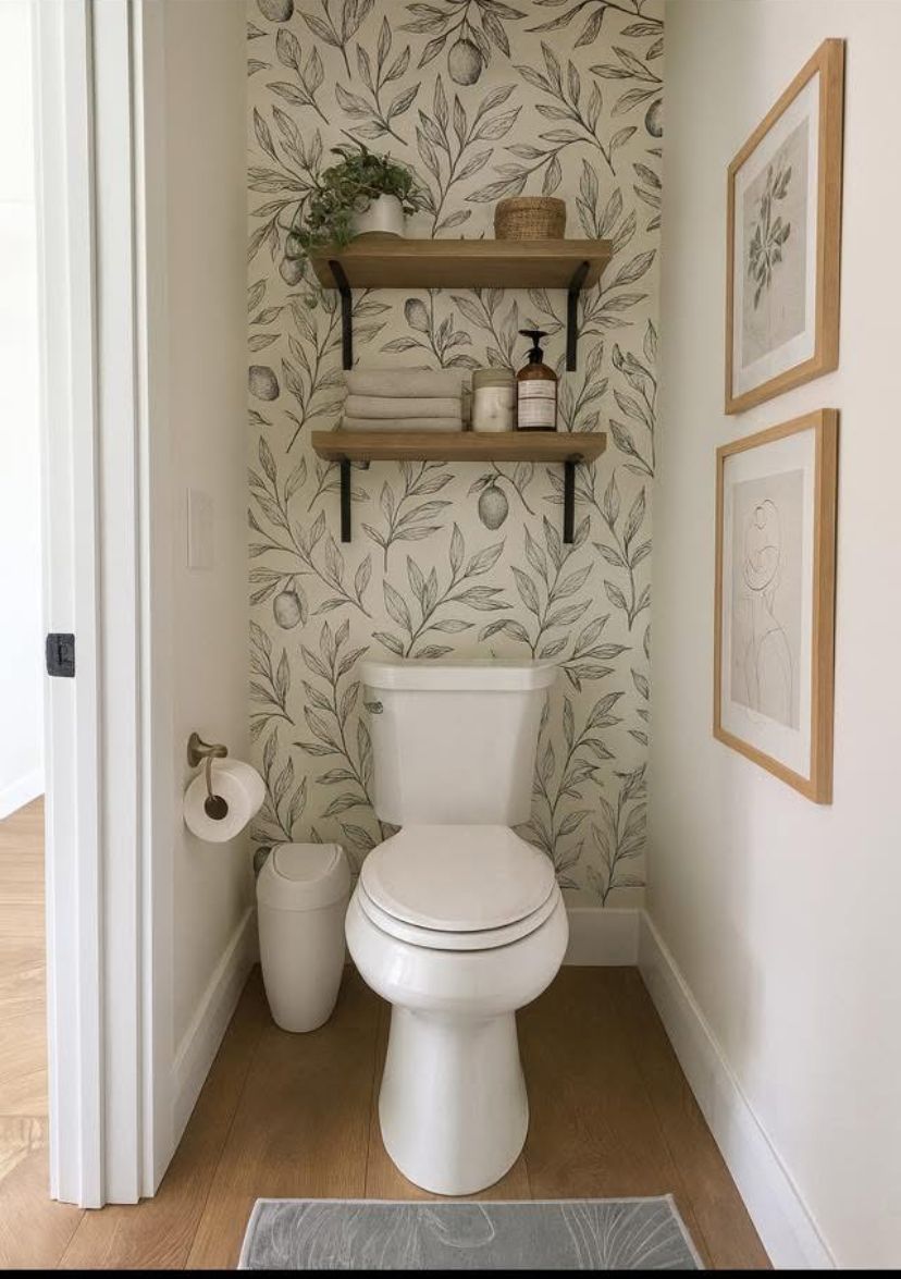 Cream leaf-print wallpaper behind two wood shelves above a toilet, styled with a plant, towels, a candle and framed line art