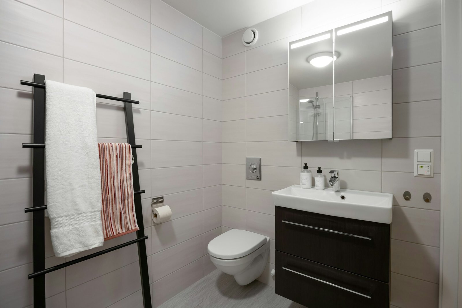 Black ladder towel rack and backlit mirrored medicine cabinet above a dark wood vanity in a small tiled bathroom