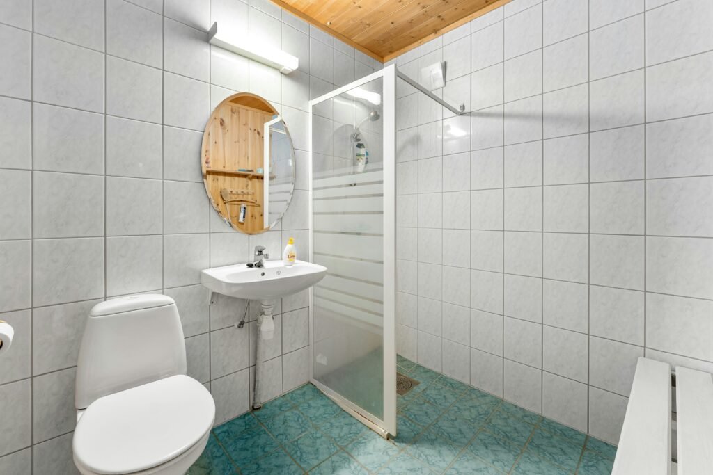Walk-in shower behind a frosted glass panel next to a round wood-framed mirror shelf in a small white-tiled bathroom with patterned teal floor tile
