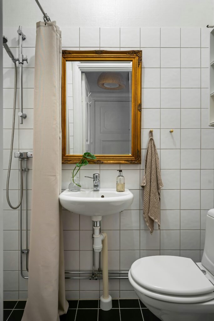 Antique gold-framed mirror above a pedestal sink with a linen hand towel, a trailing plant and a canvas shower curtain in a white tiled bathroom