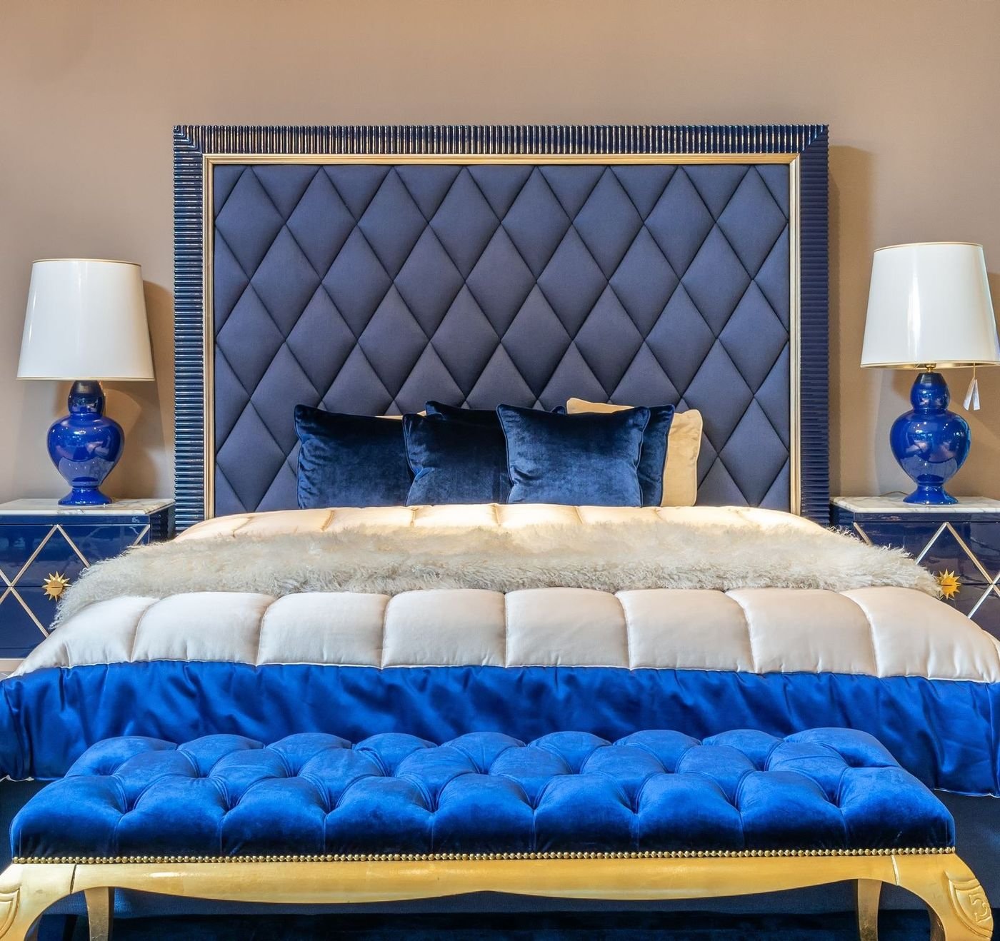 Glam bedroom with a tufted navy velvet channel-back headboard, a royal blue velvet bench with gold trim, mirrored nightstands and cobalt ceramic lamps