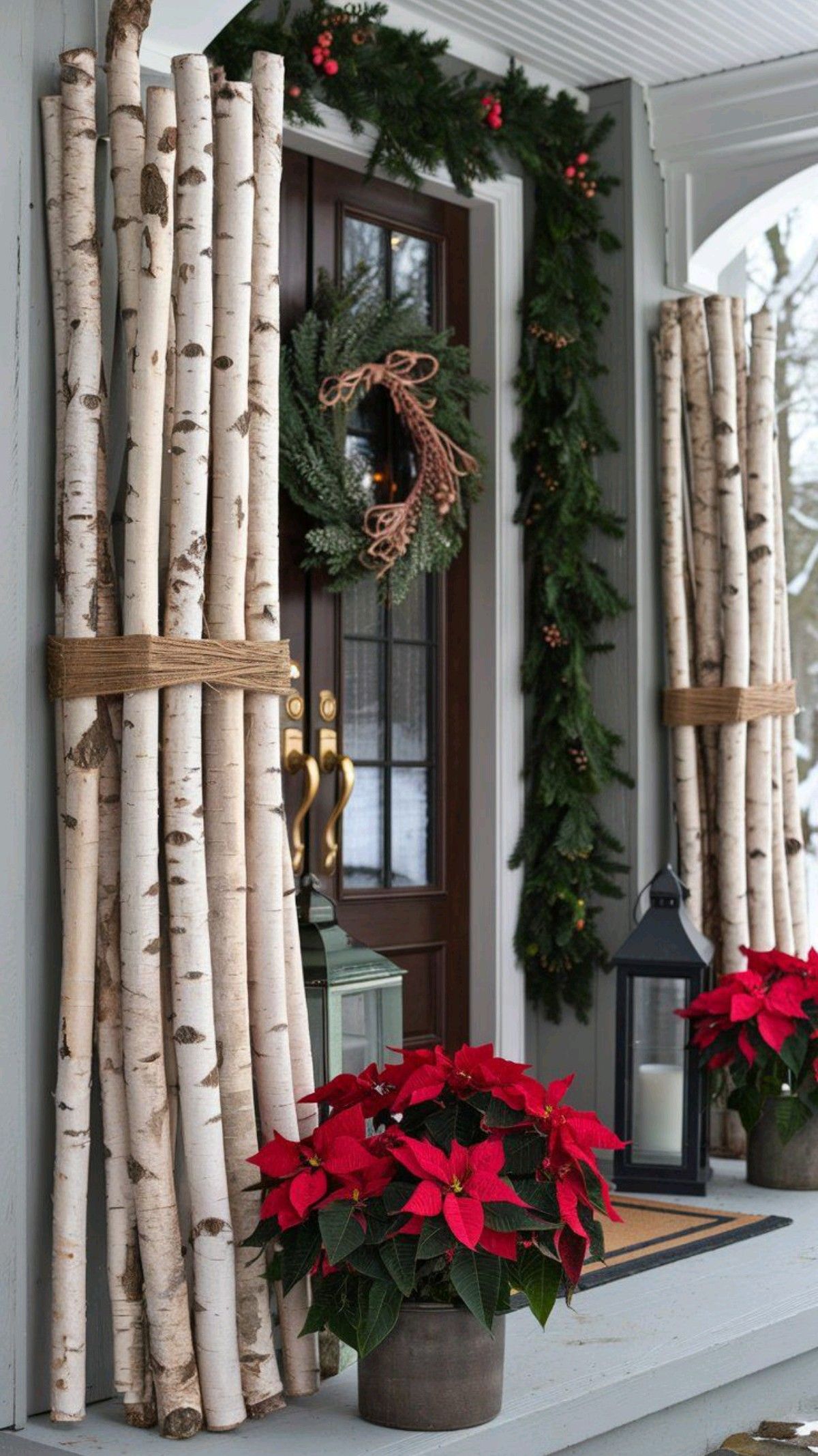Bundles of birch branches tied with twine flanking a front door with garland, a wreath, lanterns and poinsettias