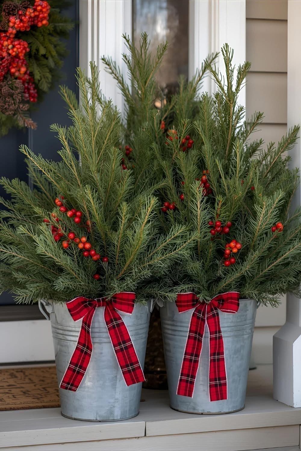 Galvanized buckets filled with evergreen branches, red berries and red plaid bows on a porch step