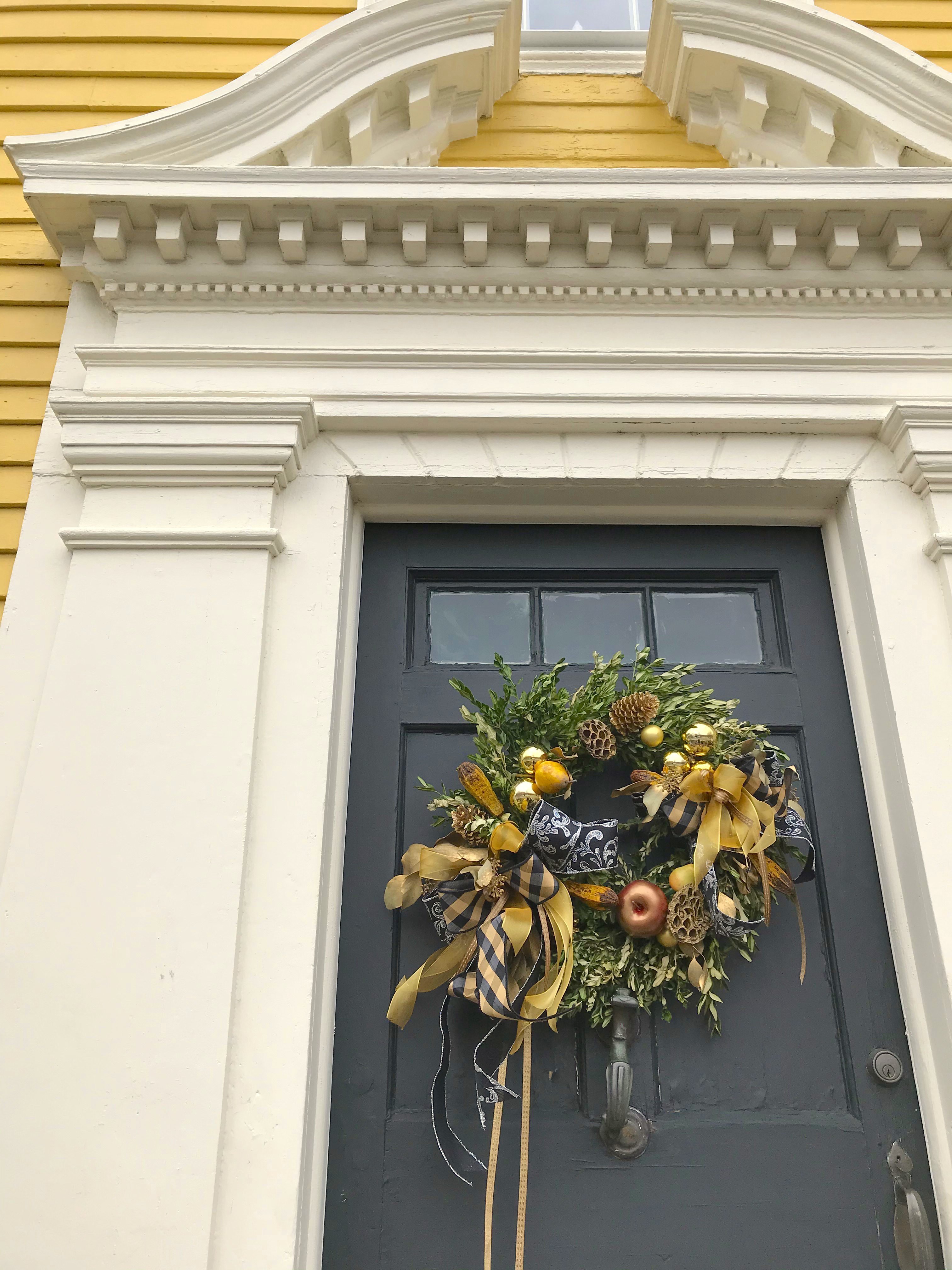A navy front door on a historic yellow house decorated with a boxwood wreath tied with plaid and gold ribbon