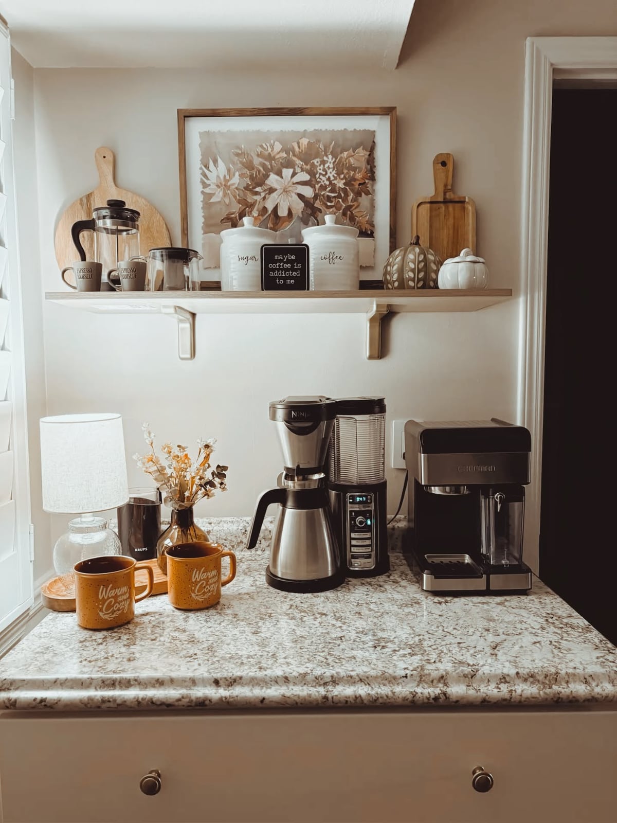 Speckled counter with two coffee makers, two mugs and a small lamp beneath a single floating shelf with framed art and canisters