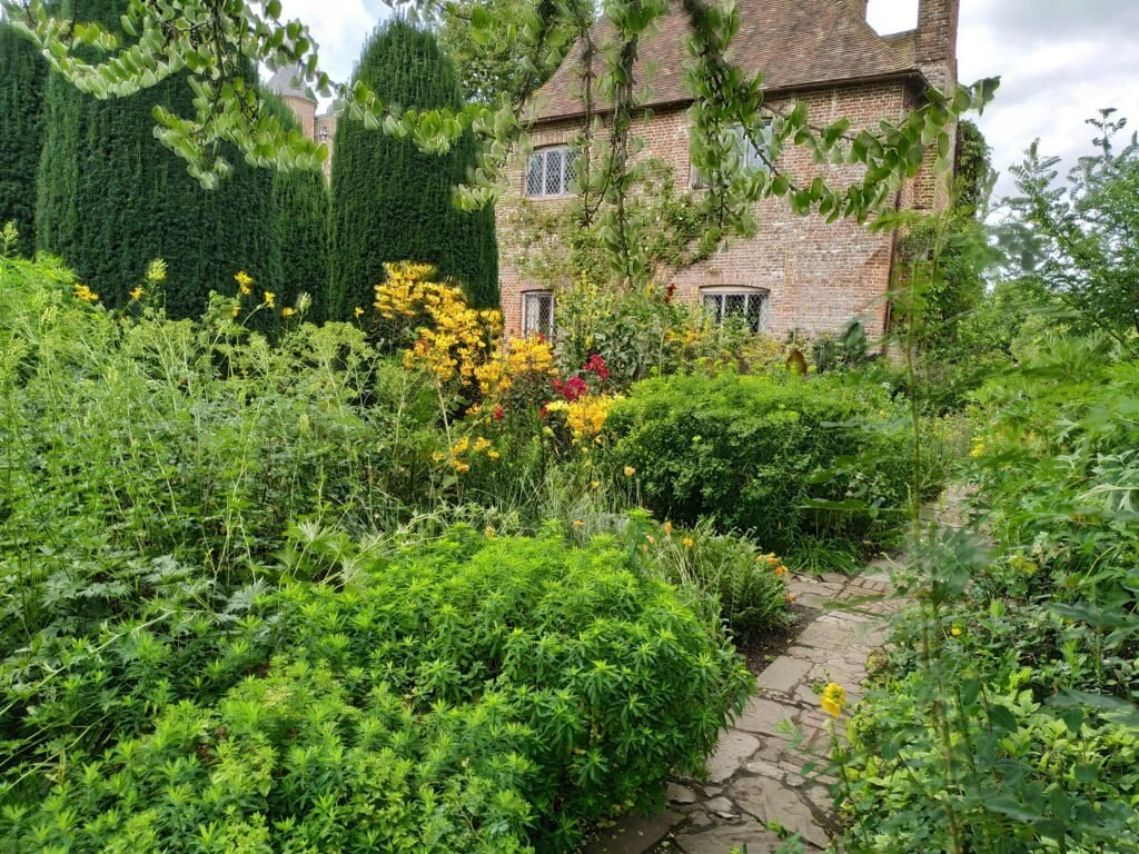 Brick cottage surrounded by dense green shrubs, climbing vines and a stone pathway leading toward the entrance