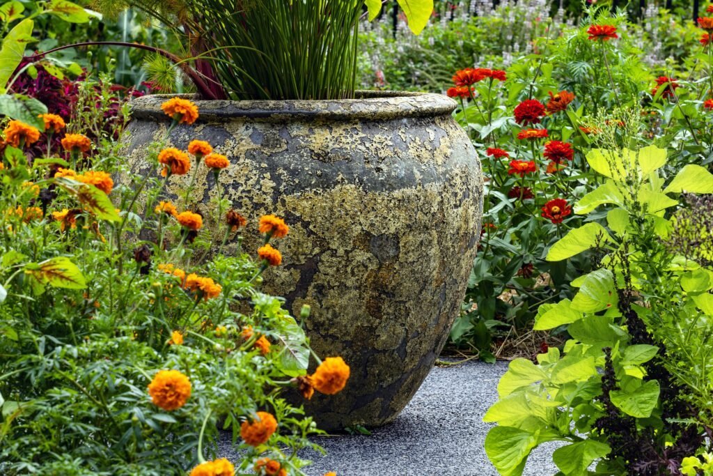 Large weathered terracotta garden urn surrounded by orange marigolds and red zinnias along a gravel path