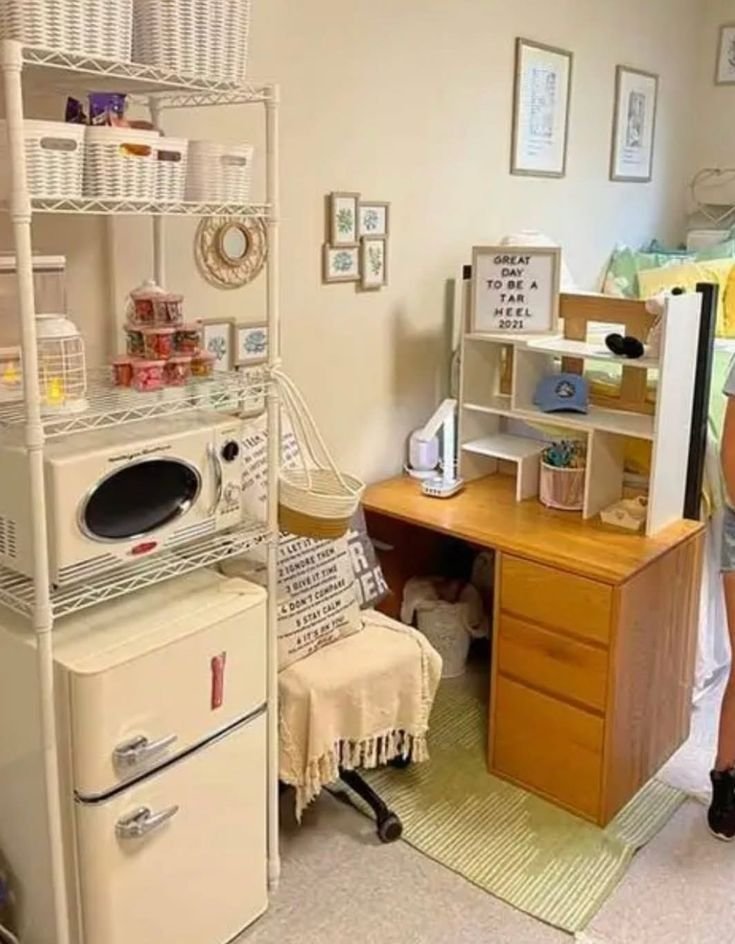 White wire shelving unit in a dorm room holding woven baskets, a microwave and a mini-fridge, next to a desk with photos and decor