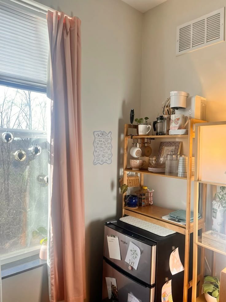 Tall wood shelving unit in a dorm room stacking a coffee maker, mugs and dishes above a black mini-fridge with a microwave on top