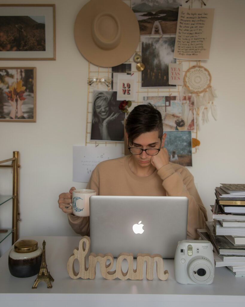 Student sitting at a desk with a laptop and mug, backed by a large personal mood board wall with photos, a straw hat, a dreamcatcher and handwritten notes