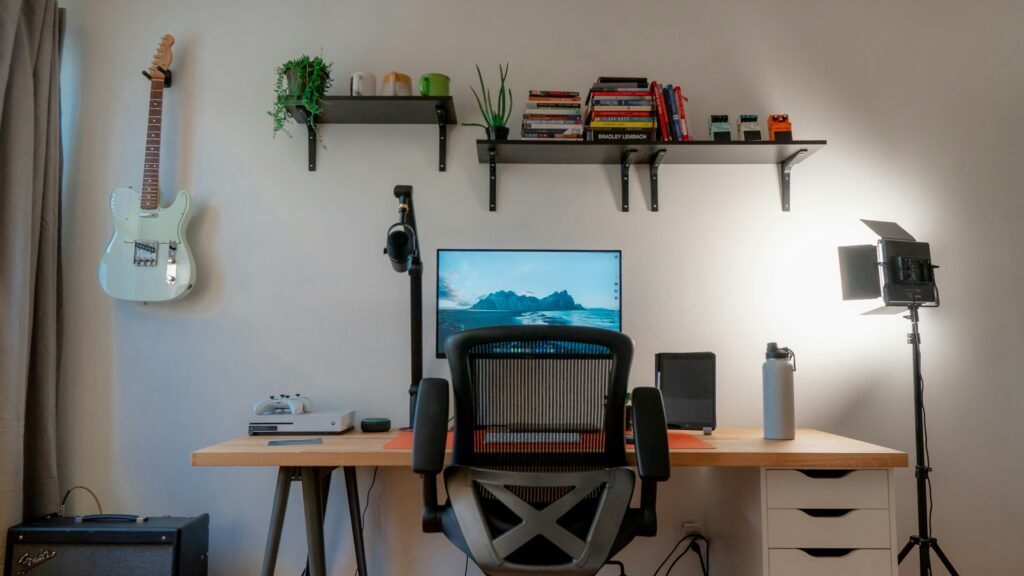 Two black floating shelves mounted above a desk holding books, small plants and mugs, with a guitar hung on the wall beside them