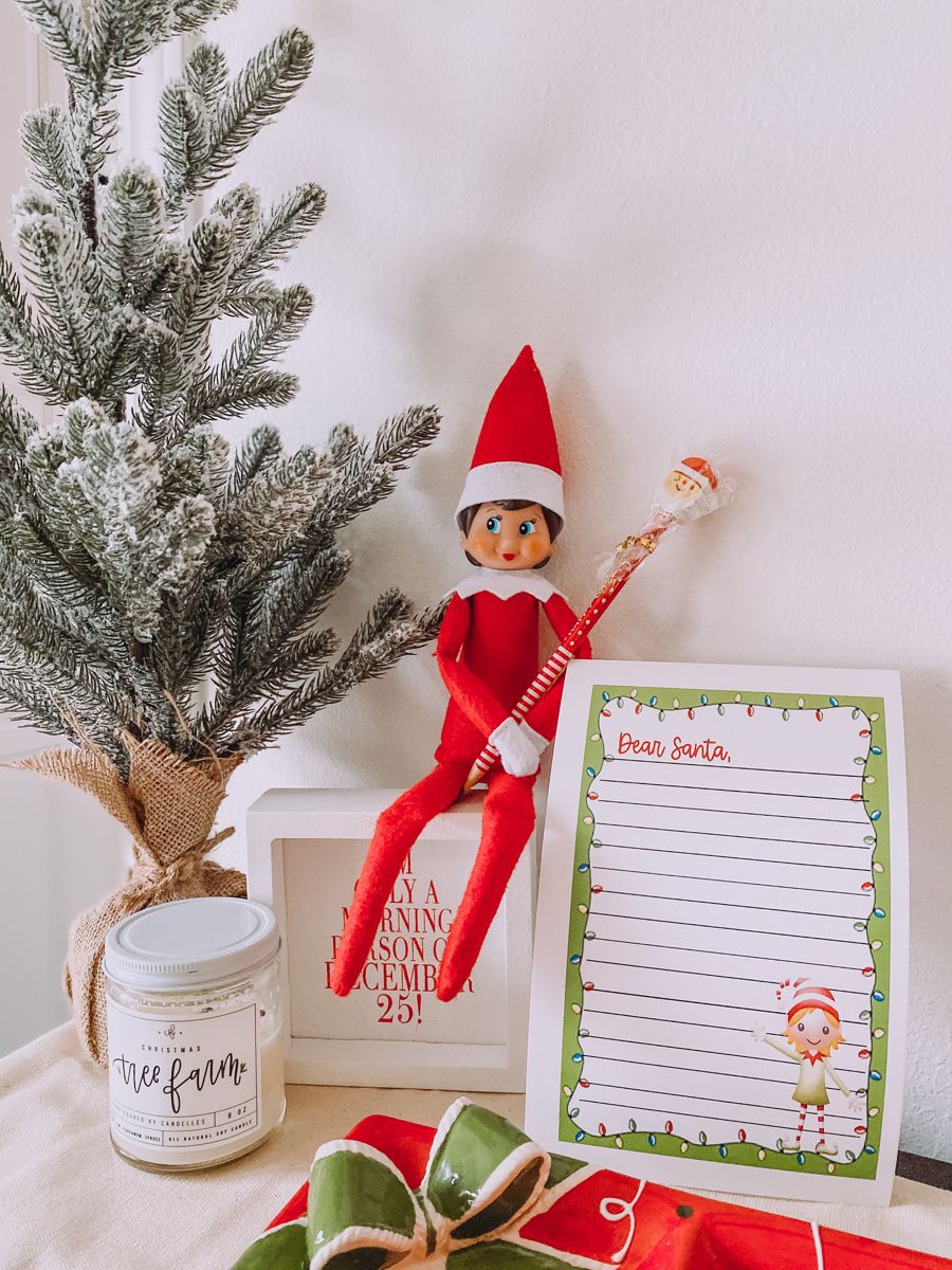 Elf on the Shelf sitting on a small table holding a candy cane pen beside a letter addressed Dear Santa