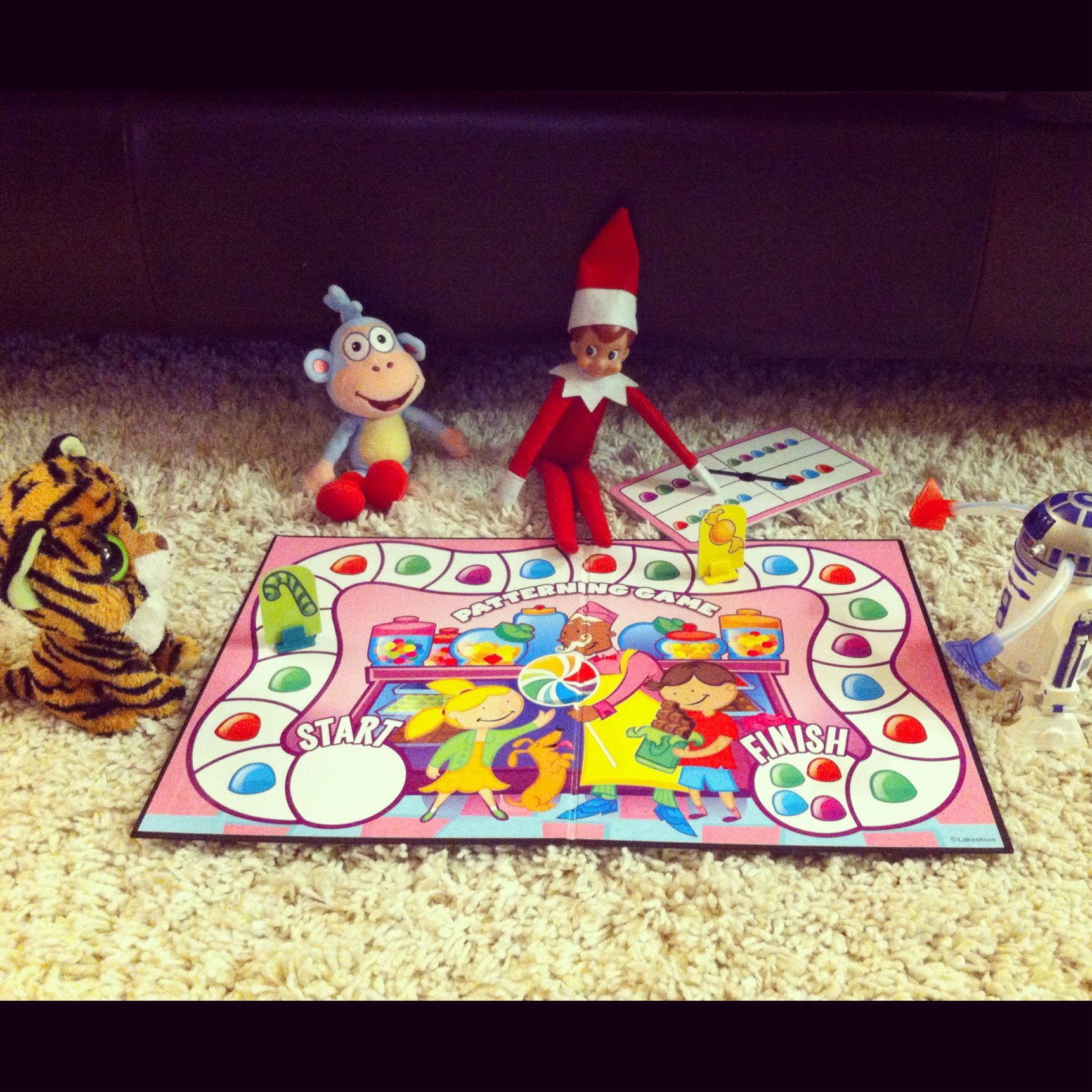Elf on the Shelf playing a childrens board game on the floor with a stuffed monkey, tiger and toy robot as friends
