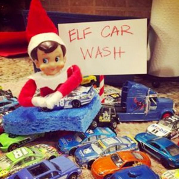 Elf on the Shelf sitting on a sponge beside a handwritten Elf Car Wash sign surrounded by toy cars