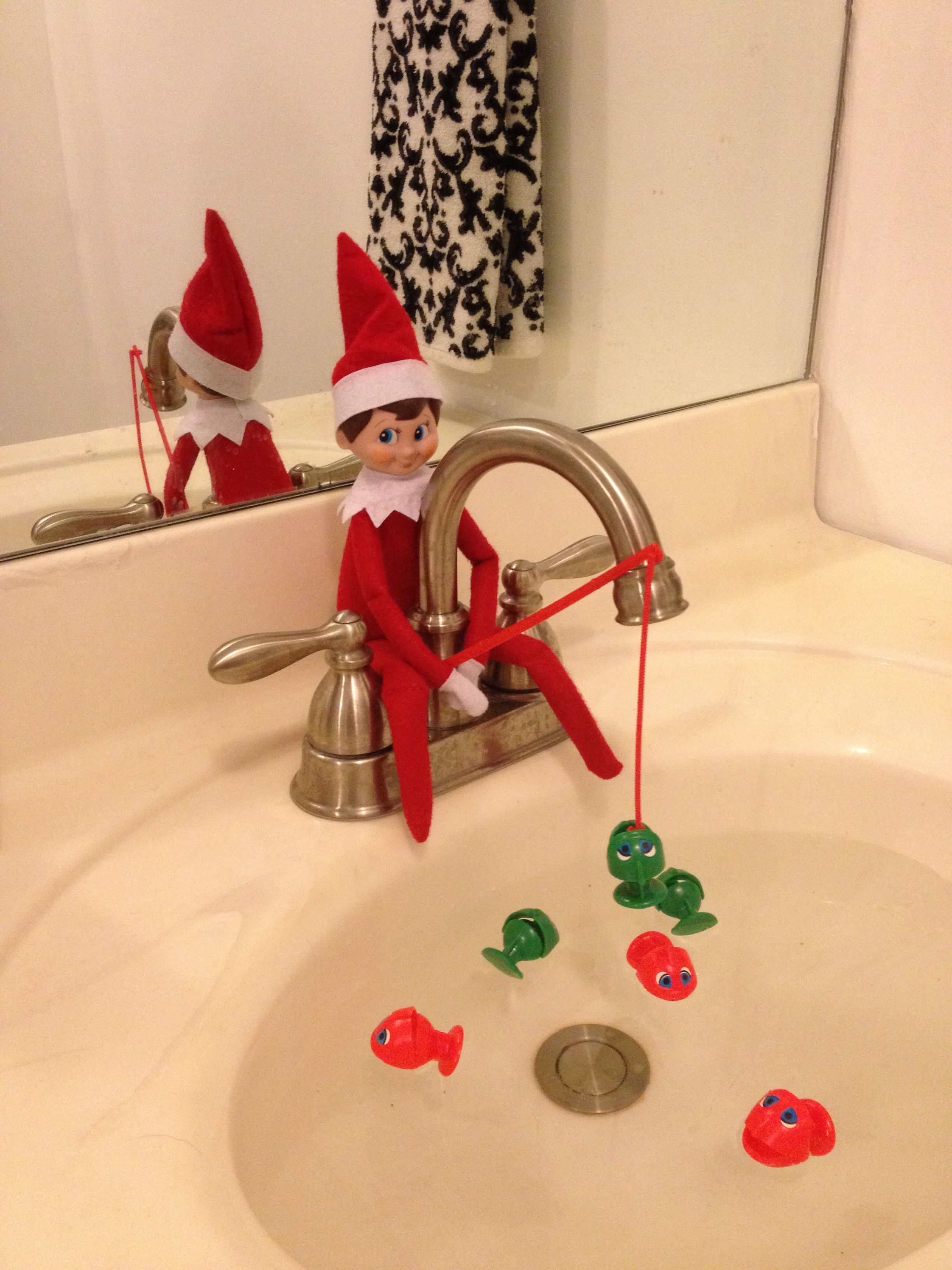 Elf on the Shelf sitting on a bathroom faucet fishing with a red string for toy fish floating in the sink