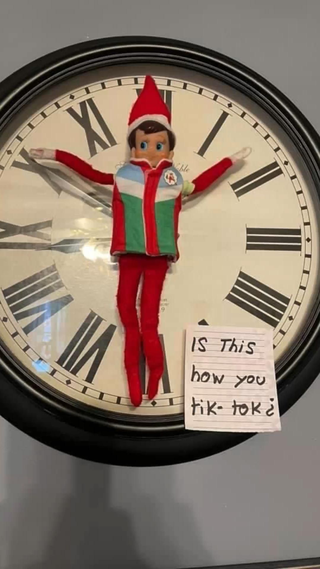 Elf on the Shelf hanging spread-eagle on a wall clock beside a handwritten note