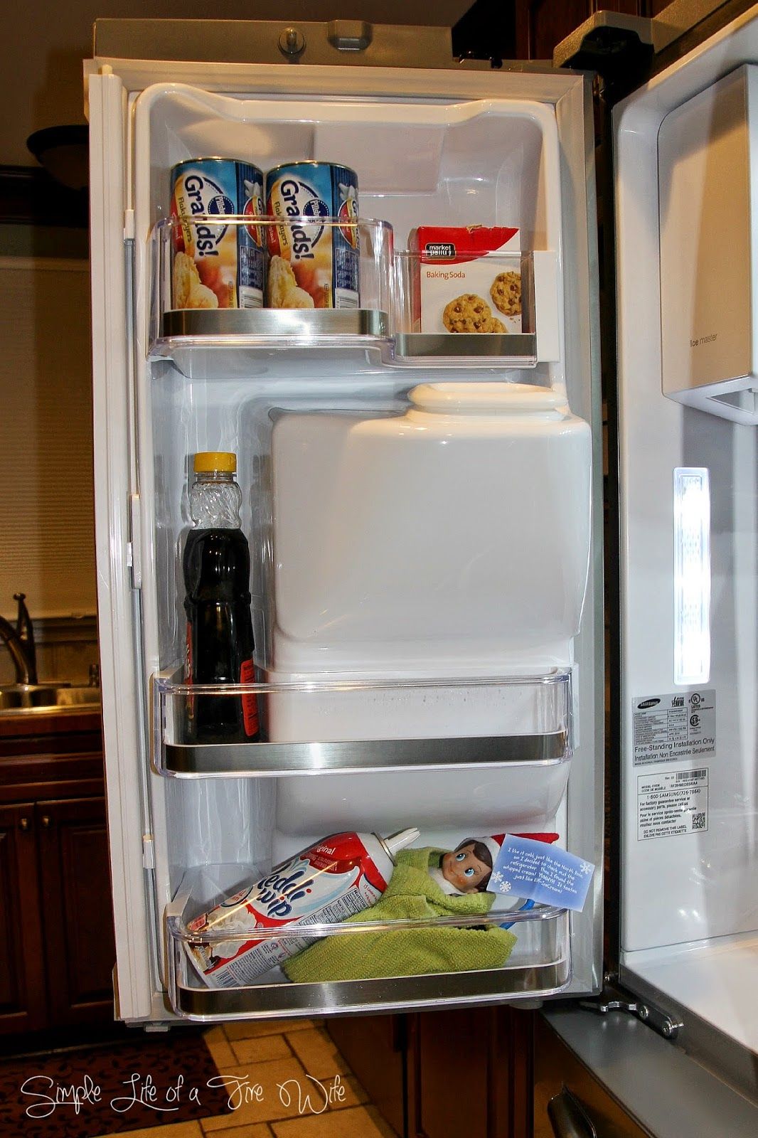 Elf on the Shelf wrapped in a green towel tucked into the door shelf of an open refrigerator beside whipped cream and condiments