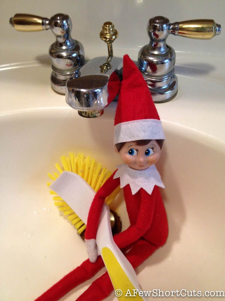 Elf on the Shelf sitting in a bathroom sink holding a scrub brush as if helping to clean