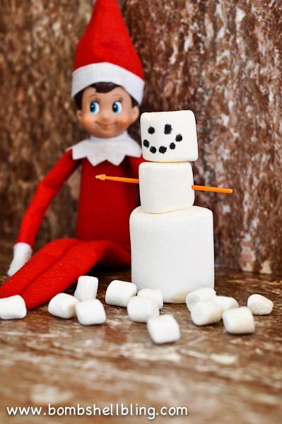 Elf on the Shelf sitting beside a small snowman built from stacked marshmallows with mini marshmallows scattered around