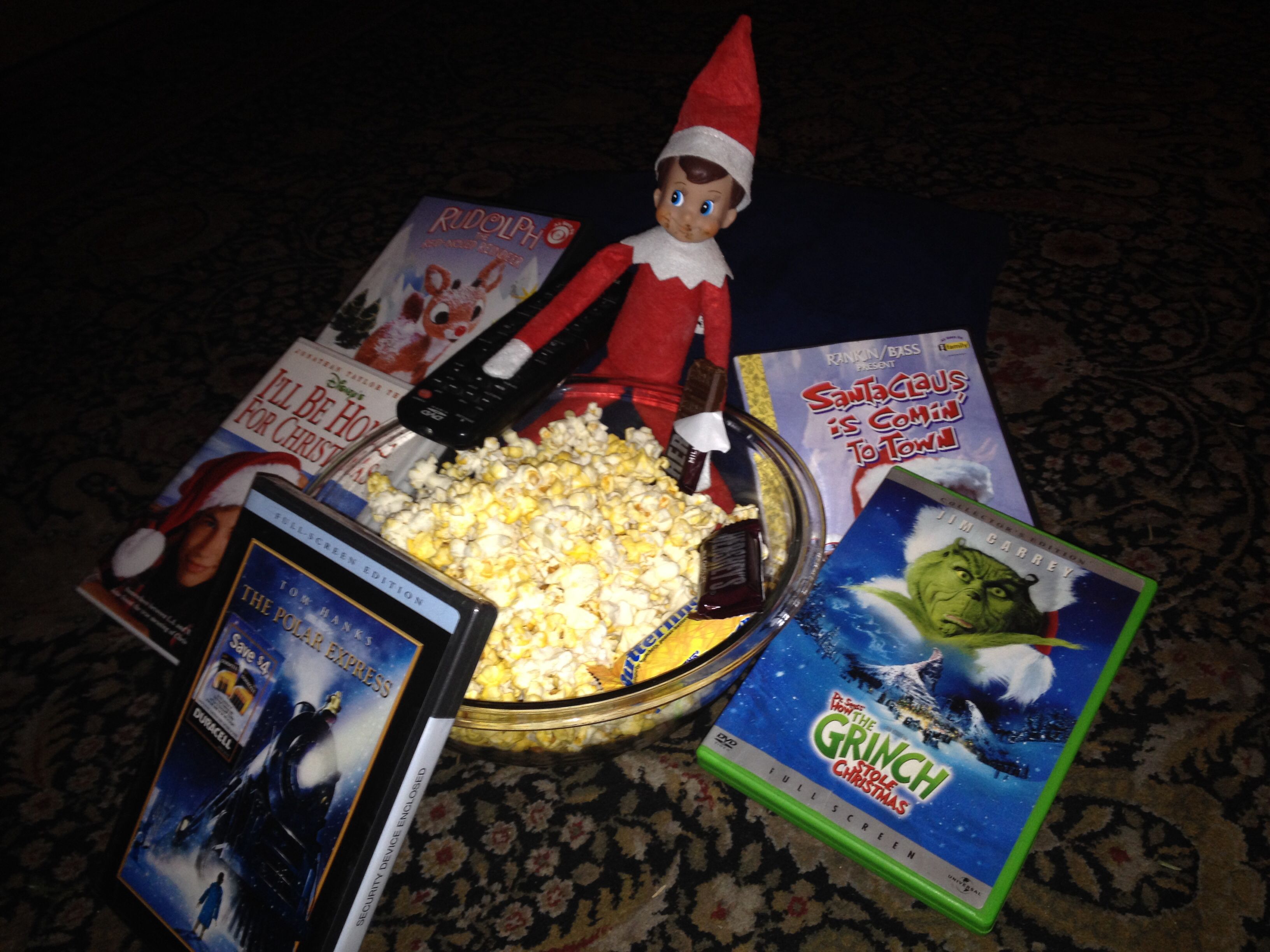 Elf on the Shelf sitting in a bowl of popcorn with a TV remote surrounded by Christmas movie DVDs