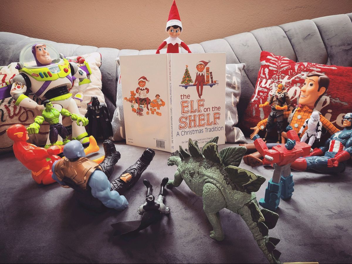 Elf on the Shelf sitting atop the Elf on the Shelf book surrounded by a circle of action figures and stuffed toys as if reading to them