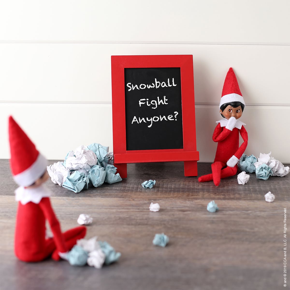 Two Elf on the Shelf dolls sitting on either side of a chalkboard sign reading Snowball Fight Anyone surrounded by balled-up paper snowballs