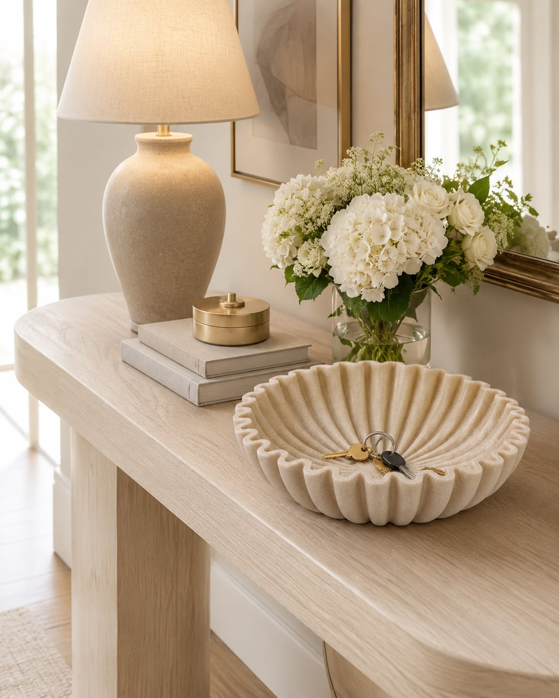 Warm table lamp glowing beside a vase of white flowers and a scalloped bowl on an entryway table