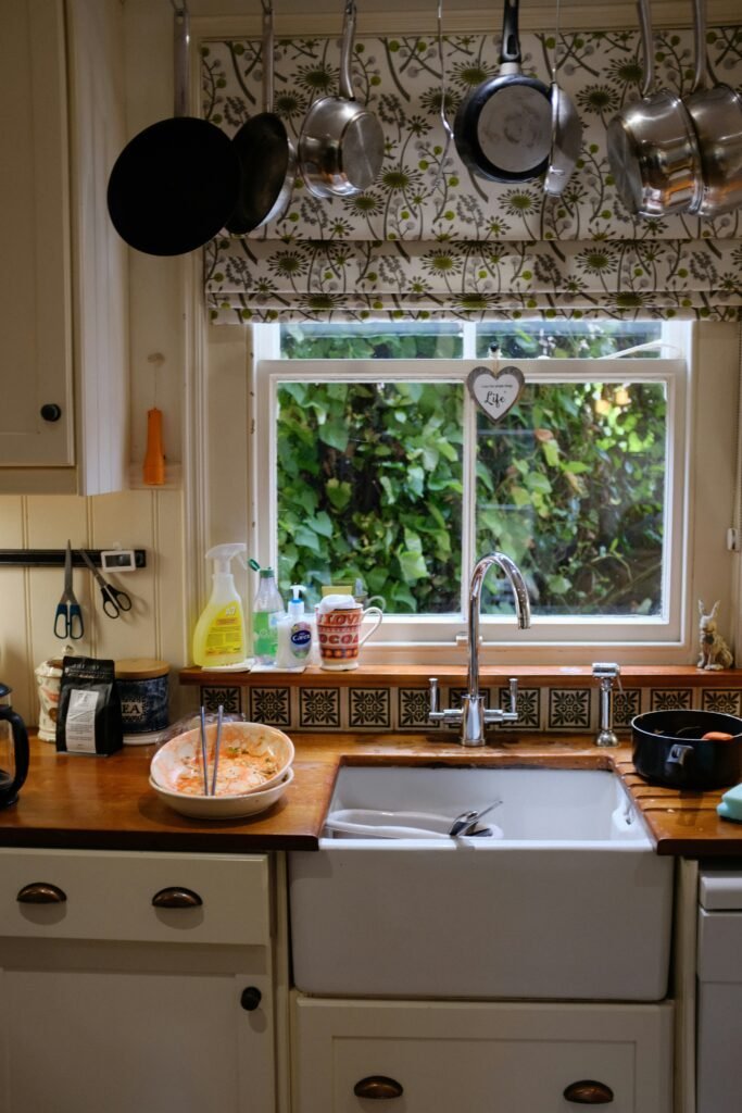 White apron-front farmhouse sink below a window with pots and pans hanging above and a floral curtain valance