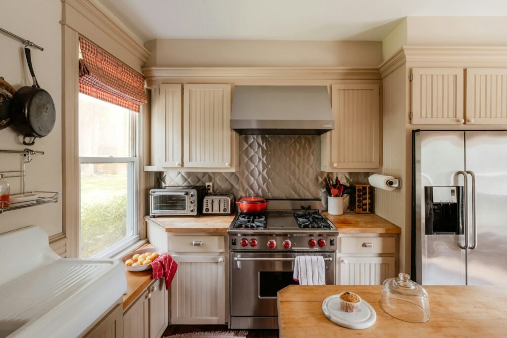 Cream beadboard kitchen cabinets with a wood butcher block counter, red gingham curtain and stainless steel range