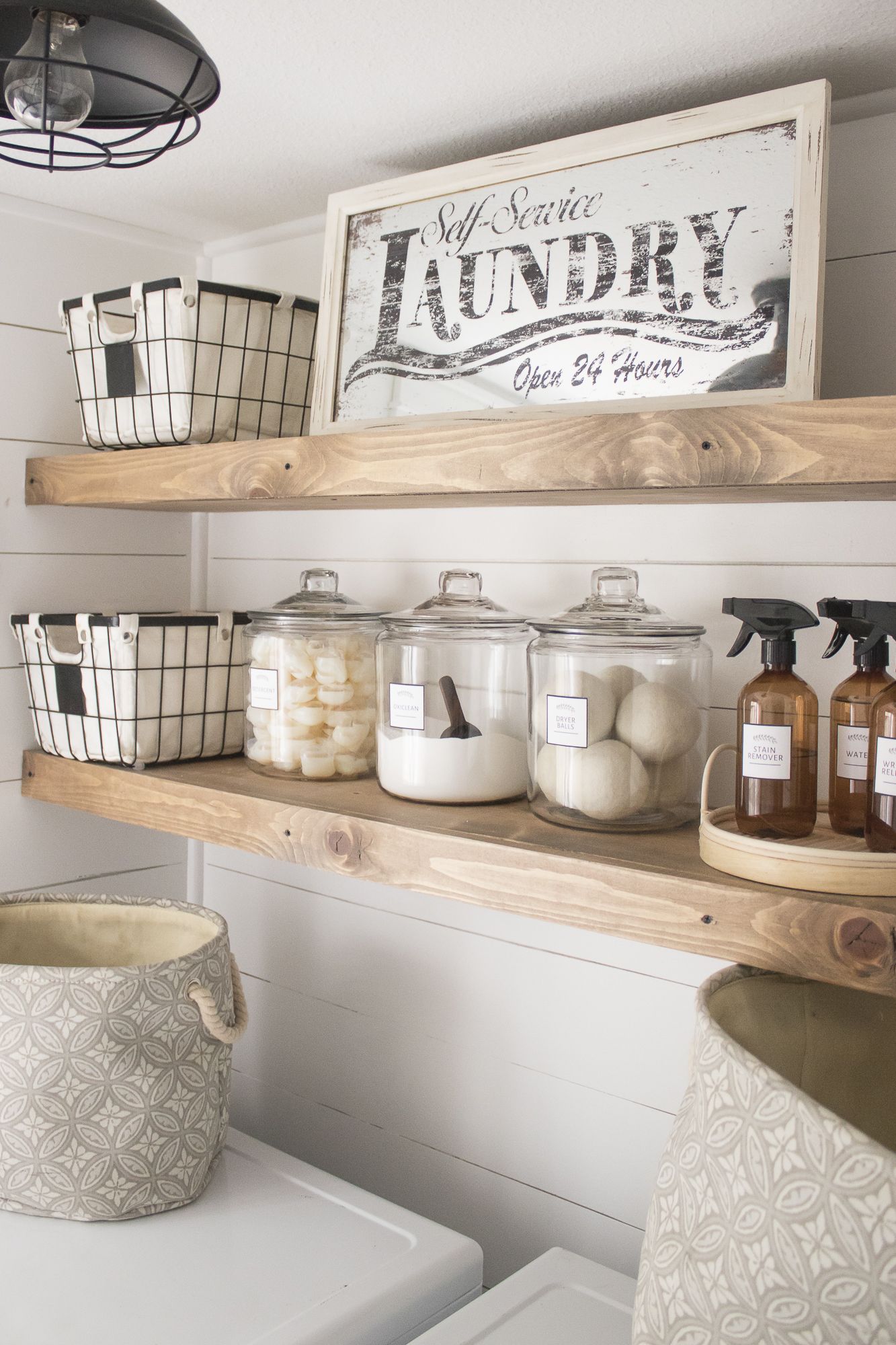 Matching glass jars labeled detergent, Oxiclean and dryer balls on a laundry room shelf with a vintage sign