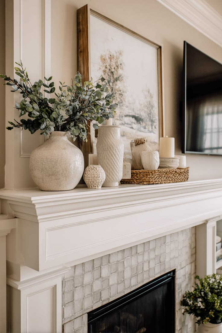 White mantel styled with textured ceramic vases, eucalyptus and a woven tray of candles beneath a leaning landscape print and mounted TV