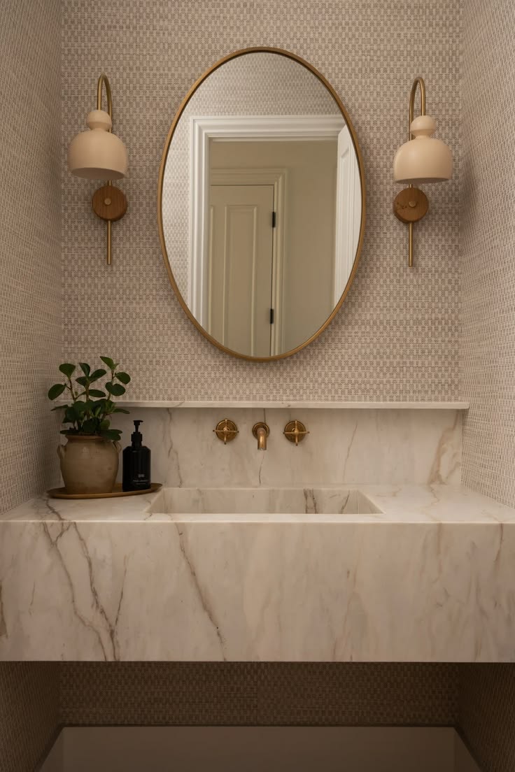 Oval brass mirror flanked by cream drum-shade sconces above a marble backsplash and vessel sink