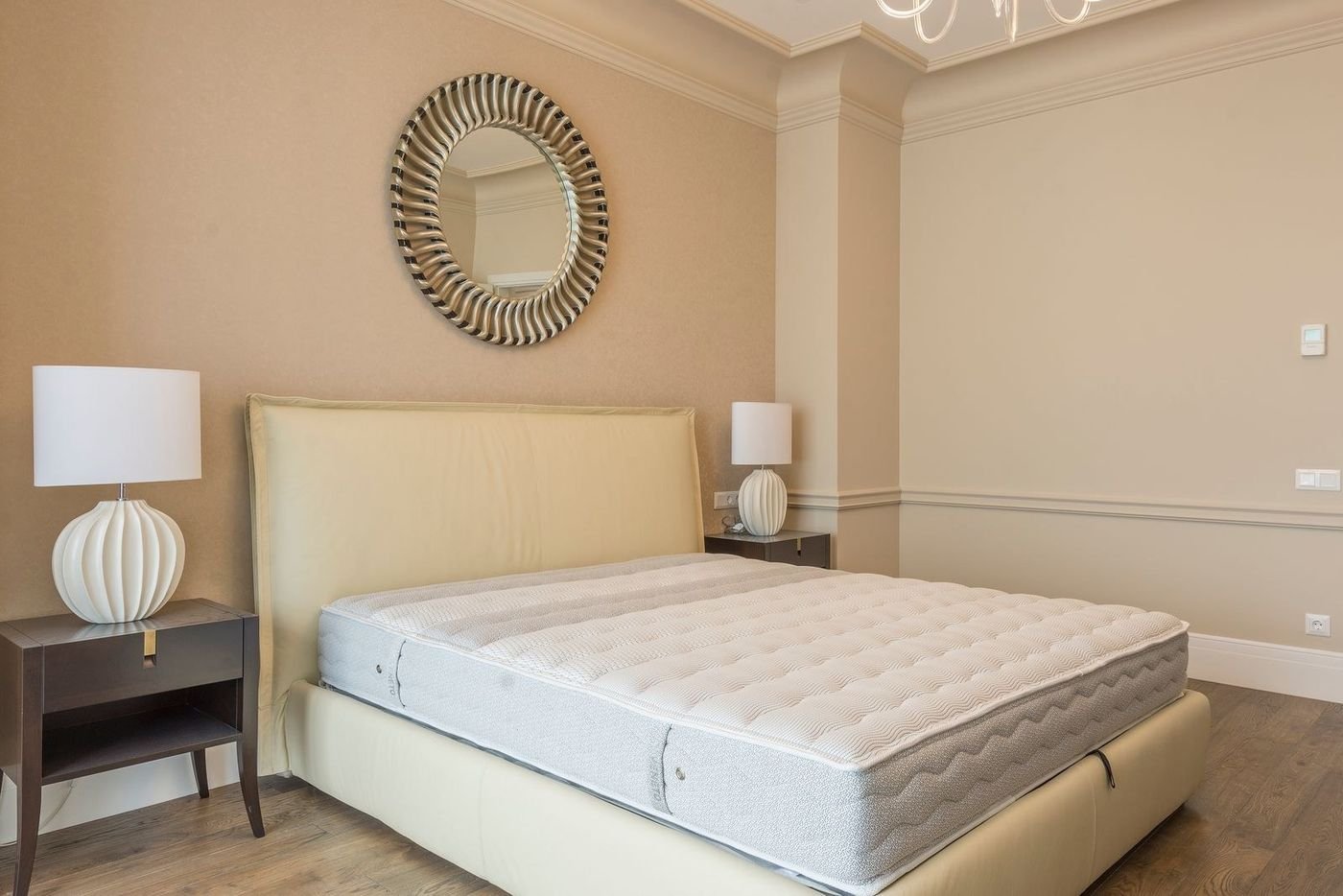Small bedroom with warm neutral walls, a round mirror above a tufted headboard and layered bedding