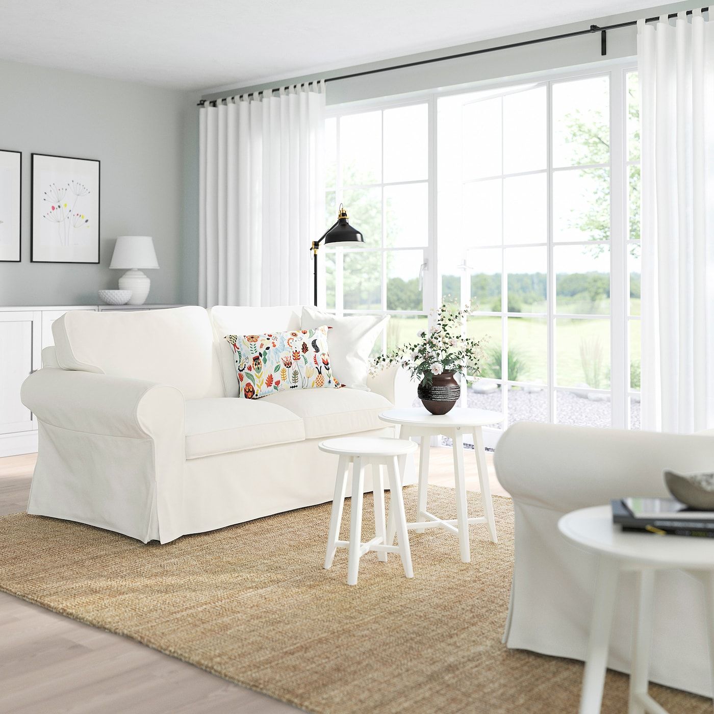 White slipcovered sofa with a floral pillow, a white armchair and two small white stools on a jute rug in a bright room