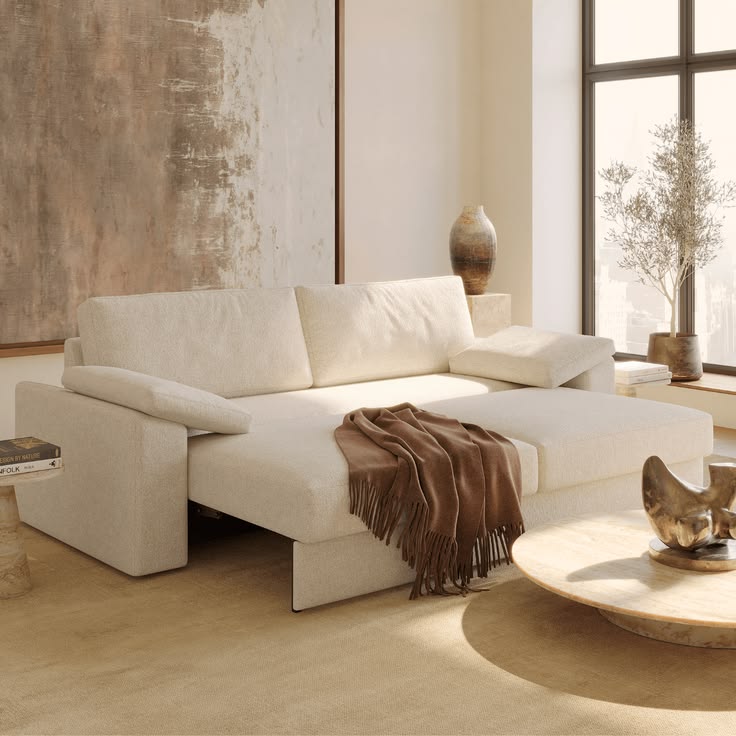Cream sofa with its seat extended forward for lounging, a brown fringed throw and a round stone coffee table