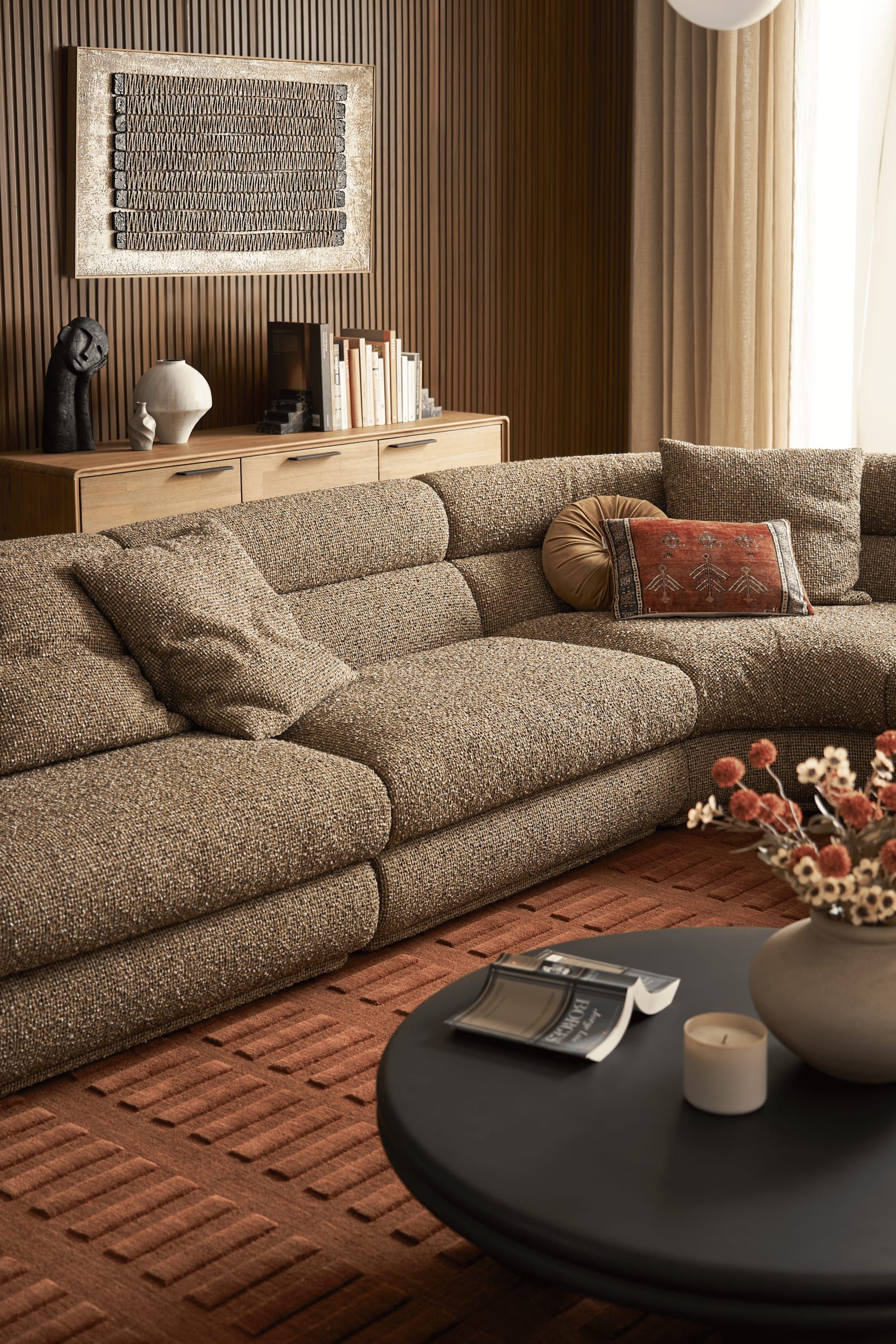 Large taupe textured sectional in front of a wood-slat wall with a rust patterned rug and a black round coffee table