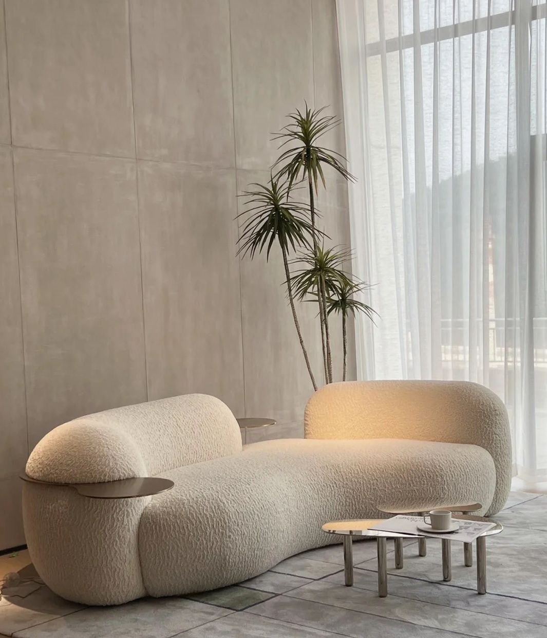 Sculptural cream bouclé sofa with a low curved back and a small metal tray on one arm, beside a tall plant