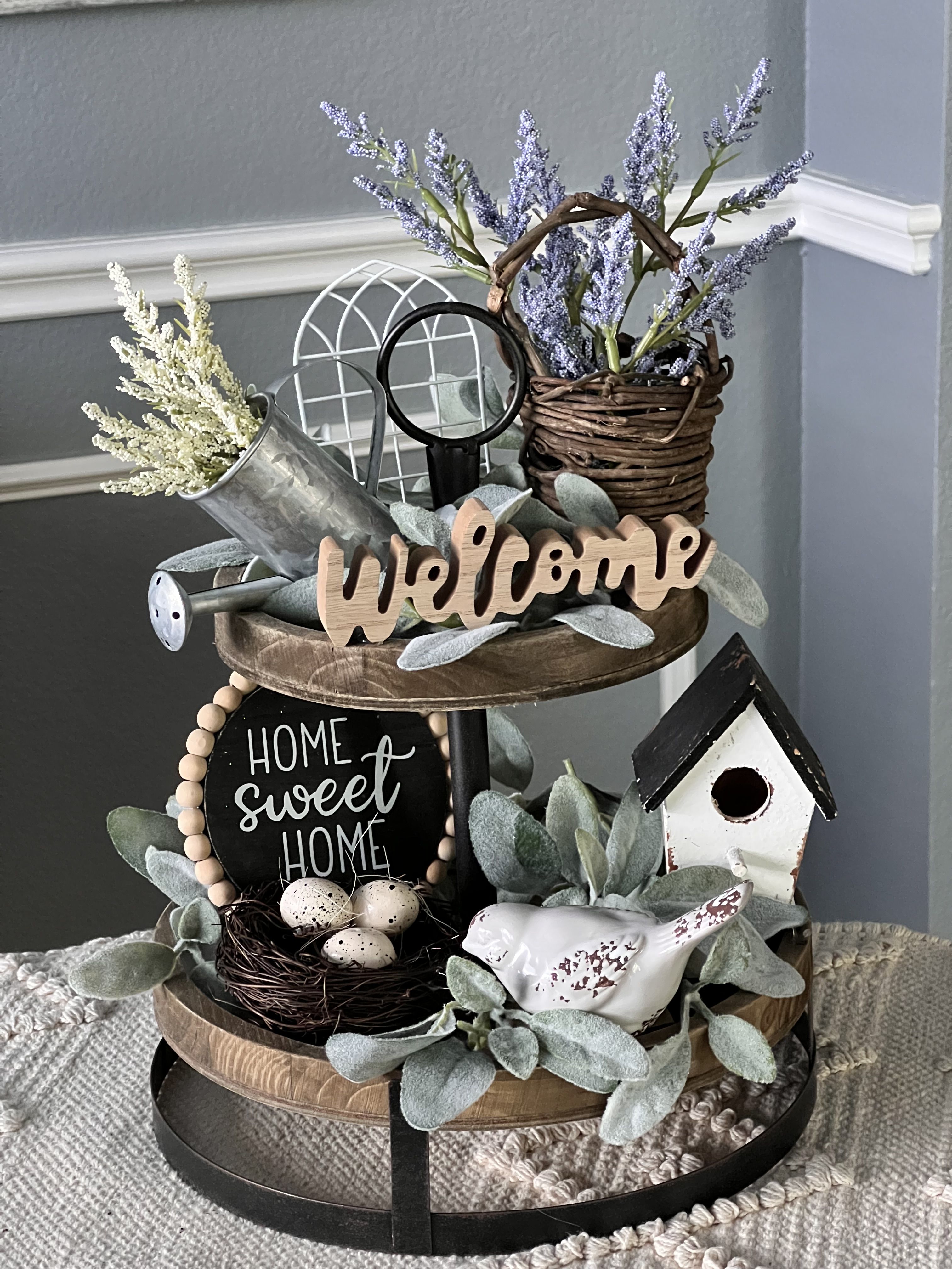 Two-tier wood and metal tray styled as an entryway display with a Welcome sign, Home Sweet Home sign, a bird nest and a birdhouse