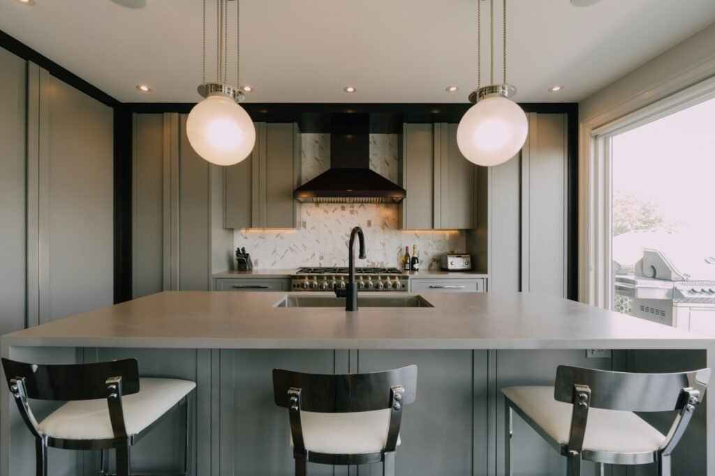 Gray island kitchen with two glass globe pendant lights, dark wood bar stools and a marble tile backsplash with a black hood