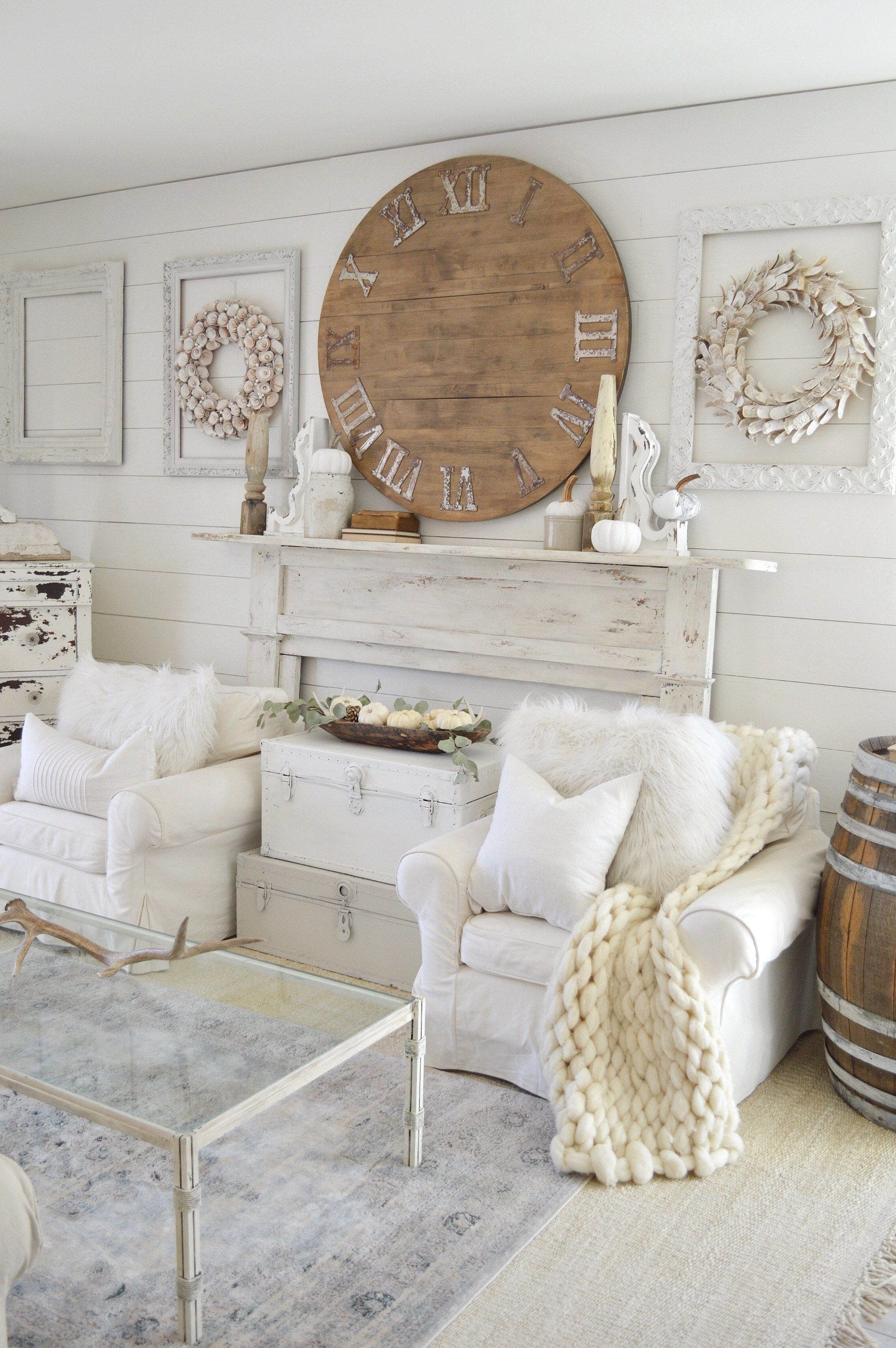 White farmhouse living room with a large round wood clock, distressed mantel, wreaths and chunky knit throws