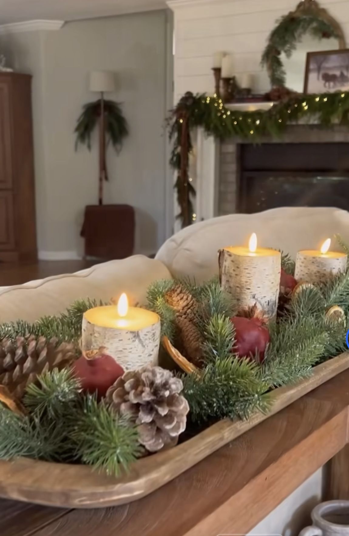 Dough bowl centerpiece with birch bark candle holders, lit candles, pinecones, pomegranates, dried citrus and evergreen on a sofa back