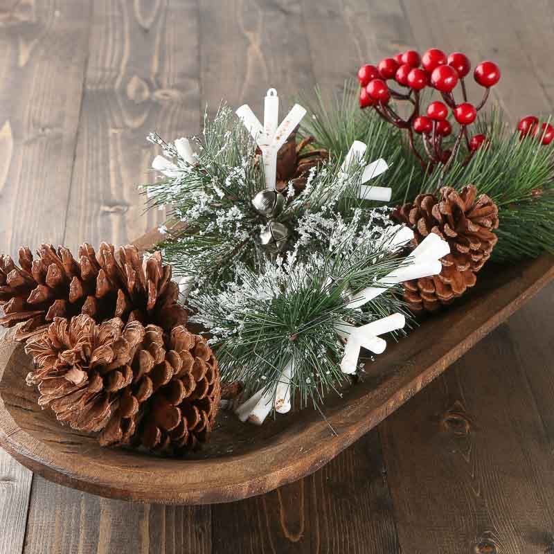 Elongated dough bowl with pinecones, frosted greenery, small silver bells and red berries on a dark wood table