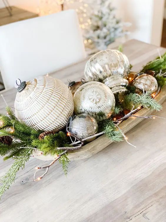 Light wood dough bowl filled with oversized textured mercury glass ornaments, fairy lights and pine branches on a dining table